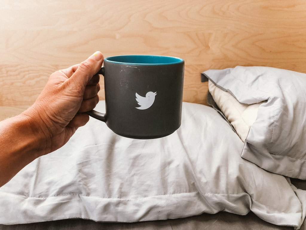 Growing on Twitter Blackhat Strategy man holding black cup with Twitter logo and white sheet bed background beige wall