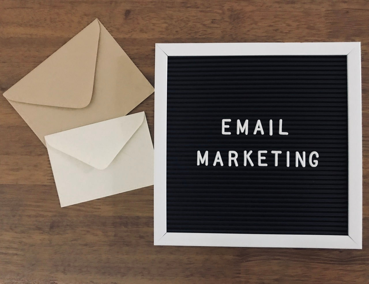 Two envelopes, one smaller white one bigger brown. Words "Email Marketing" written in all capital letters on square blackboard with white frame.